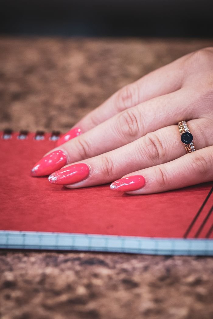 Close-up of a hand with pink manicured nails and a ring resting on a notebook.