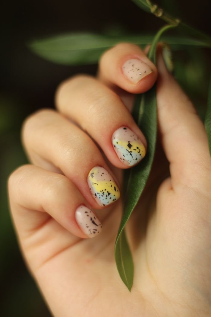 Artistic nail design with colorful speckles, surrounded by green leaves for a natural touch.
